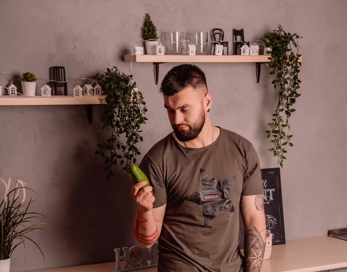 Homme barbu examinant un concombre dans une cuisine stylée avec étagères.